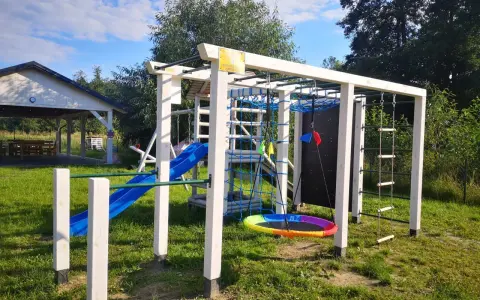 Plac zabaw w **dolinie roztoczy**: zjeżdżalnia, drabinki, huśtawka i ścianka wspinaczkowa. W tle altana.