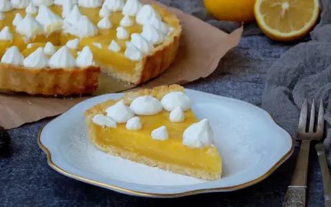 Une part de tarte au citron, garnie de meringue, sur une assiette. La tarte entière est visible en arrière-plan. Une recette de tarte au citron parfaite pour le dessert.
