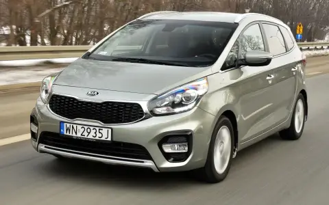 Srebrny Kia Carens, typowy MPV, jedzie po drodze. To auto rodzinne, idealne na wycieczki.