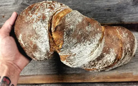 Ein rustikales wurzelbrot mit sauerteig, bestäubt mit Mehl, liegt auf einem Holzbrett.