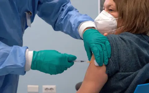 Osoba w niebieskim fartuchu i zielonych rękawiczkach podaje zastrzyk. Kobieta w masce KN95 otrzymuje trzecią dawkę szczepionki.