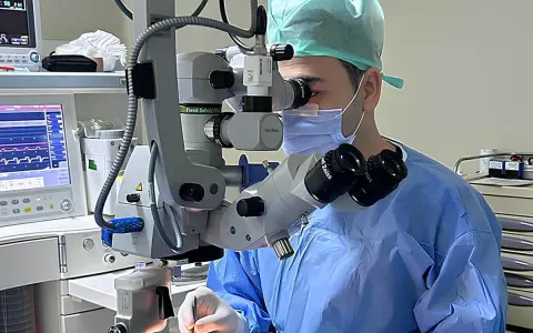 Chirurgo con maschera e camice blu opera con microscopio Zeiss. Monitor vitale e strumentazione indicano un intervento post-operatorio vitrectomia gas.