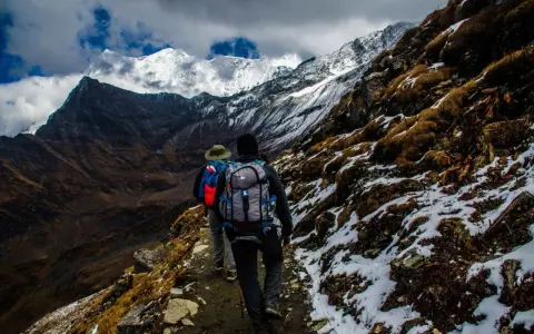 Dwóch turystów z plecakami wędruje ścieżką u podnóża ośnieżonych gór, przypominających krajobraz Mount Kenya.