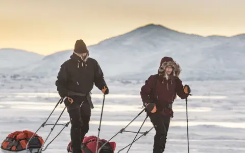 Zwei Personen ziehen Schlitten durch verschneite Landschaft. Die Berge im Hintergrund sind mit Schnee bedeckt. Ein Abenteuer wie aus dem Fjällräven g-1000 Katalog.