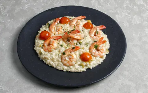 Kremowe risotto z krewetkami, pomidorkami koktajlowymi i posiekaną natką pietruszki na czarnym talerzu.