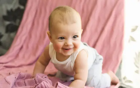 Ein lächelndes Baby krabbelt auf einer rosafarbenen Decke. Es ist etwa 26 Wochen alt und erlebt gerade einen Entwicklungsschub.