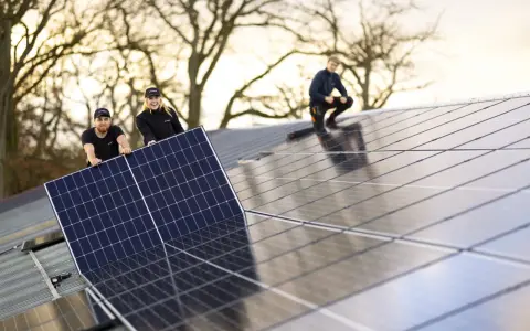 Arbetare installerar solpaneler på ett tak. En av dem, en kvinna, ler glatt medan hon håller i en panel. En annan man hjälper till, och en tredje person sitter på taket. De är en del av teamet för **solverkets alfa**.
