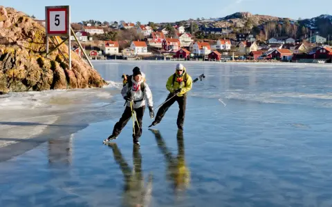 Två personer åker skridskor på en isbelagd sjö, med en pittoresk by i bakgrunden. De använder stavar för att hjälpa sig fram, vilket visar hur åker man skridskor på ett effektivt sätt.