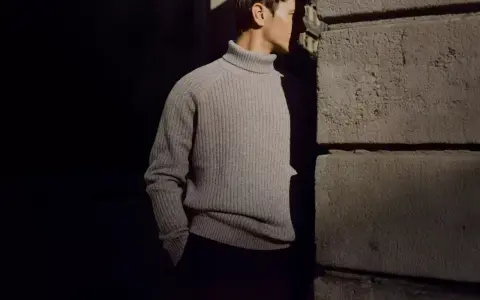 Uomo con un elegante outfit maglione collo alto grigio, in piedi vicino a un muro di pietra.