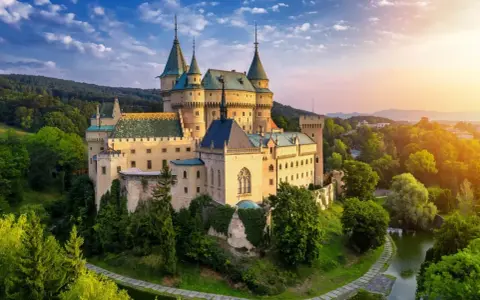 Bajkowy zamek otoczony zielenią, idealne miejsce na nocleg w zamku. Słońce zachodzi nad wzgórzami, tworząc magiczną atmosferę.