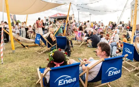 Ludzie odpoczywają na leżakach z logo Enea podczas festiwalu. W tle scena i tłum.