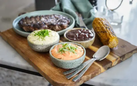 Grillad biff med tre olika såser till grillat: en vit med örter, en mörk och en orange. Majskolv och bestick ligger på en träbricka.