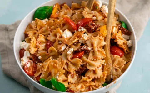 Ein köstlicher, sommerlicher Nudelsalat mit Farfalle, Tomaten, Feta und Basilikum, serviert in einer weißen Schüssel mit Holzutensilien.