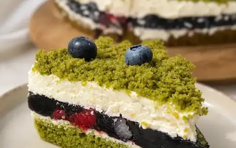 Kawałek tortu z zielonym biszkoptem, kremem, galaretką i owocami, posypany okruszkami przypominającymi leśny mech.