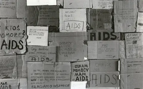 Złe ojciec, zniszczony przez AIDS, błaga o pomoc. Kartony z napisami "AIDS", "SOS", "Ratunku!" i prośbami o wsparcie.