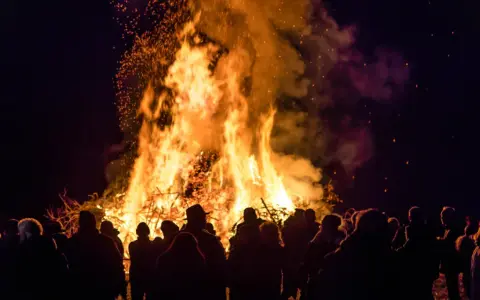 Menschen versammeln sich um ein helles Osterfeuer. Die Flammen lodern hoch in den Nachthimmel, Funken fliegen. Das Osterfeuer symbolisiert die christliche Bedeutung von Licht und Auferstehung.