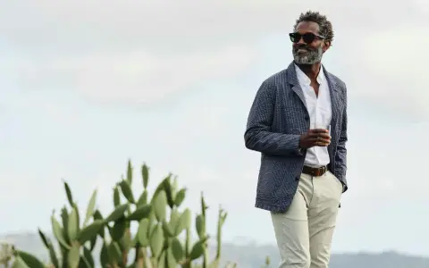 Man in stylish beach wedding attire, wearing a seersucker jacket, white shirt, and light pants, holding a drink.