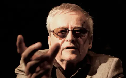 Jerzy Jarocki w okularach, z gestem dłoni, w ciemnym tle.