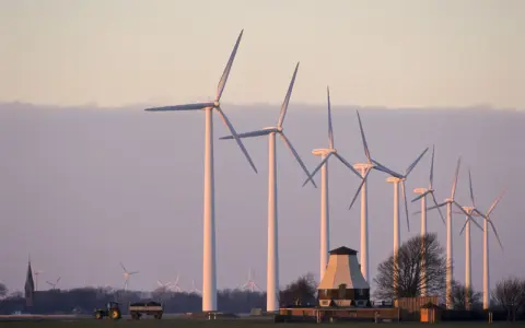 Windräder im Abendlicht, ein Traktor mit Anhänger und ein alter Mühlenbau symbolisieren die Direktvermarktung von Windstrom in ländlicher Idylle.