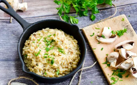 Risotto z grzybami w czarnej patelni, ozdobione natką pietruszki, obok pokrojone pieczarki na desce.