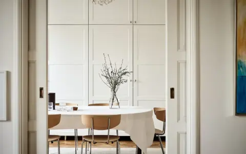 Comedor con mesa cubierta de mantel blanco, sillas de madera y un gran armario empotrado. El estilo ncs blanco aporta luminosidad.