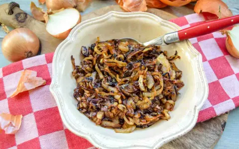 Schüssel mit karamellisierten Zwiebeln, die auf einem karierten Tuch liegen. Ganze und halbierte Zwiebeln sind daneben zu sehen.