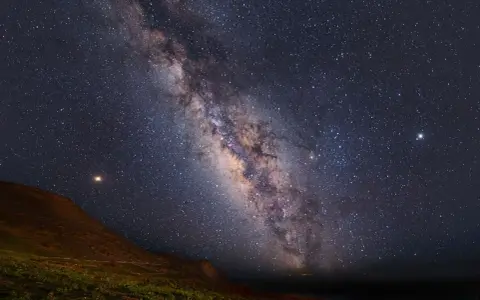 Paisaje nocturno en una reserva starlight, con la Vía Láctea cruzando el cielo estrellado sobre una colina y vegetación.