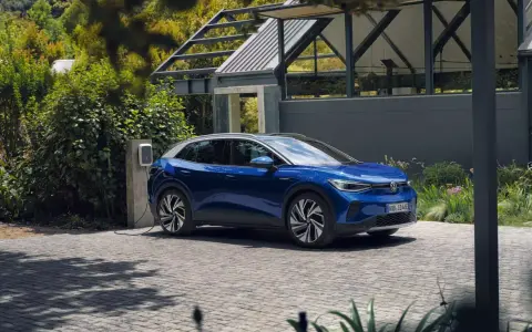 Niebieski Volkswagen ID.4 ładuje się przy domu, otoczony zielenią.
