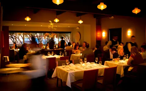 Ciemne, nastrojowe wnętrze restauracji. Goście jedzą przy stołach, kelnerzy krążą. Złe oświetlenie zdjęć jedzenia może zniechęcić gości do publikacji w social mediach.