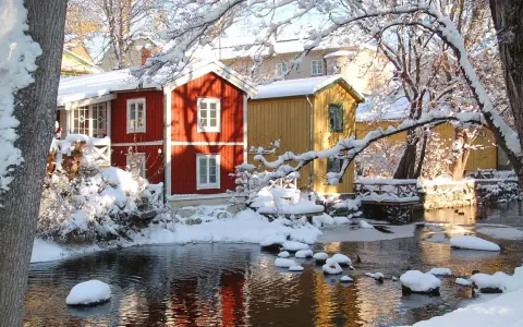Vinteridyll vid vattnet i destination Gåsvik. Röda och gula hus speglas i det isiga vattnet, omgivna av snötyngda träd.