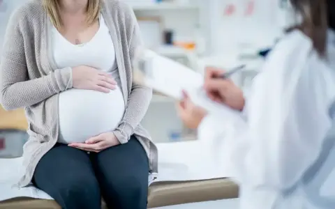 Kobieta w ciąży siedzi na kozetce, trzymając dłonie na brzuchu. Lekarz spisuje dokumenty, prawdopodobnie wystawiając zaświadczenie o ciąży.