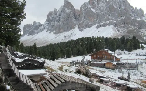 Panorami innevati con il rifugio delle Odle in lontananza, panchine in legno e pini.