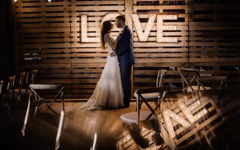 Para nietypowe sesje ślubne, para młoda pozuje przed podświetlanym napisem "LOVE" na tle drewnianych palet.