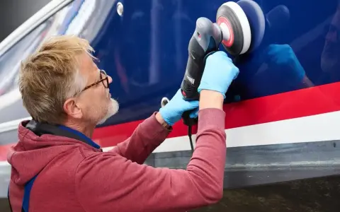 Mann poliert mit Maschine den blauen Bootsrumpf. Das Gelcoat erstrahlt nach dem Polieren wieder in neuem Glanz.