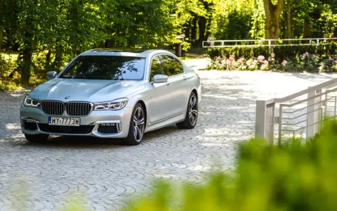 Srebrne BMW 750i lśni w słońcu na brukowanym podjeździe, otoczone zielenią i różami.