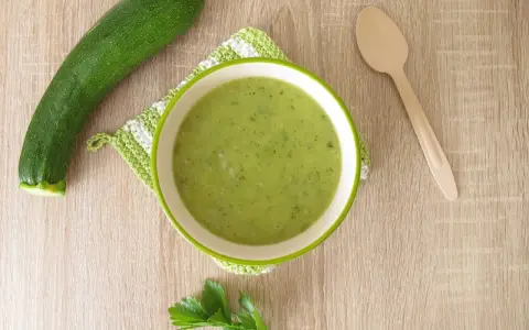 Zucchini Brei Baby: Eine Schüssel grüner Zucchinipüree, daneben eine Zucchini und ein Holzlöffel.