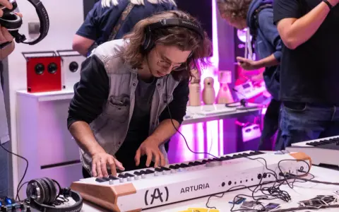 Młody mężczyzna w okularach i słuchawkach ćwiczy na keyboardzie Arturia. To część kursu produkcji muzycznej, gdzie można poznać tajniki tworzenia dźwięków.