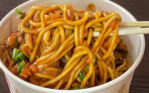 Un tazón lleno de fideos chinos salteados con verduras y salsa. Palillos levantan un bocado.