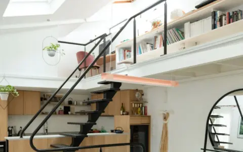 Aménagement mezzanine ouverte avec escalier métallique, cuisine en bois, étagères remplies de livres et plantes suspendues.