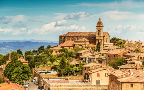 Panoramica del centro storico di Montalcino, con la chiesa e le case in pietra sotto un cielo azzurro con nuvole.