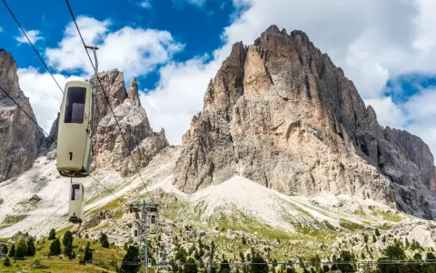 Cabine della funivia salgono verso le imponenti vette del Sassolungo, un anello di roccia maestoso sotto un cielo azzurro con nuvole bianche.
