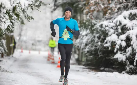 Mann läuft im Winter durch verschneiten Park. Er trägt ein blaues Shirt mit einer Startnummer in Form eines Weihnachtsbaums.