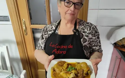 Una mujer sonriente muestra un plato de manitas de cerdo en salsa. Lleva un delantal negro con el texto "La Cocina de Adora".