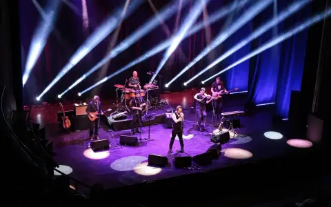 Zespół muzyczny na scenie, oświetlony niebieskimi i fioletowymi światłami. Wokalista na pierwszym planie, gitarzyści, basista, skrzypek i perkusista. Jak liczyć takty w muzyce, można się nauczyć, obserwując ich rytm.