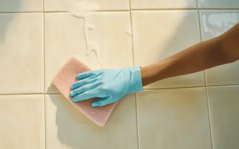 Main tenant, une main gantée de bleu frotte le carrelage de la salle de bain avec une éponge rose, faisant mousser le savon.