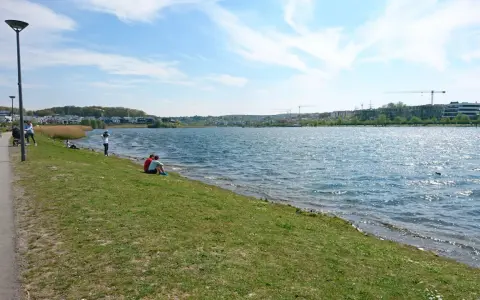 Ein sonniger Tag am Dortmunder See. Menschen spazieren am Ufer entlang, während ein Paar auf dem Gras sitzt und die Aussicht genießt.
