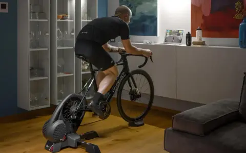 Mężczyzna trenuje na rowerze stacjonarnym z napędem **direct drive**, śledząc wirtualną trasę na laptopie.