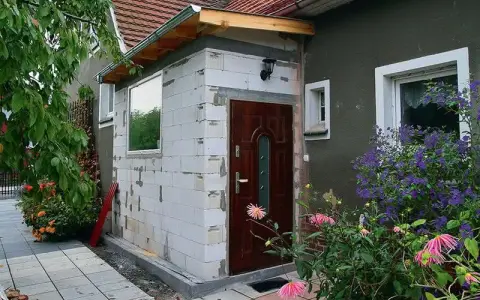 Budowa dobudowy wiatrołapu z bloczków, z nowymi oknami i drzwiami, otoczona zielenią.