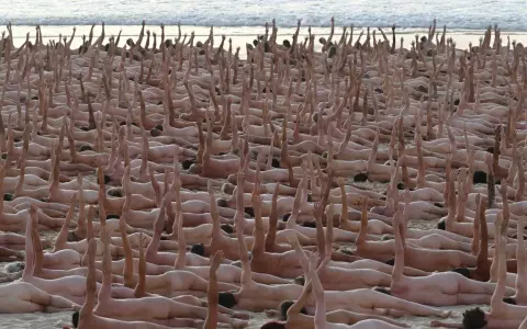 En stor grupp människor ligger på en strand, många med armarna uppsträckta. En naken målning av mänskligheten.