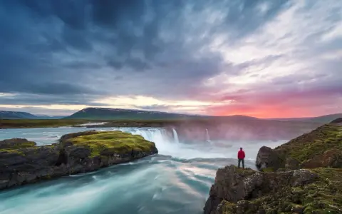 Człowiek w czerwonej kurtce podziwia wodospad na Islandii. Czy Islandia jest bezpieczna? Tak, to piękny i bezpieczny kraj.